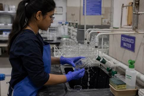 Indian researcher rinsing laboratory glassware under deionised water tap in a Karnataka university chemistry lab — apron and gloves on