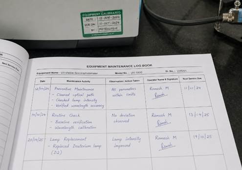Laboratory equipment maintenance logbook open on a bench in a Karnataka research lab — handwritten maintenance entries and calibration sticker on instrument behind